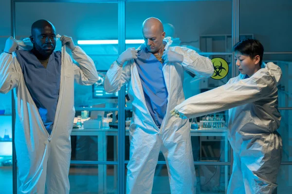 Three intercultural colleagues putting on protective coveralls in front of chemical laboratory before going to dangerous area