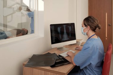 Young female radiologist in mask sitting by workplace in front of computer and looking at x-ray image of patient lungs or thorax