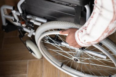 Above view of hand of young man with disability rotating wheel of wheelchair while moving down wooden floor