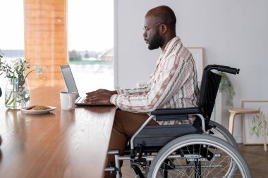 Side view of serious black man working in the net or analyzing online data while sitting in front of laptop by table