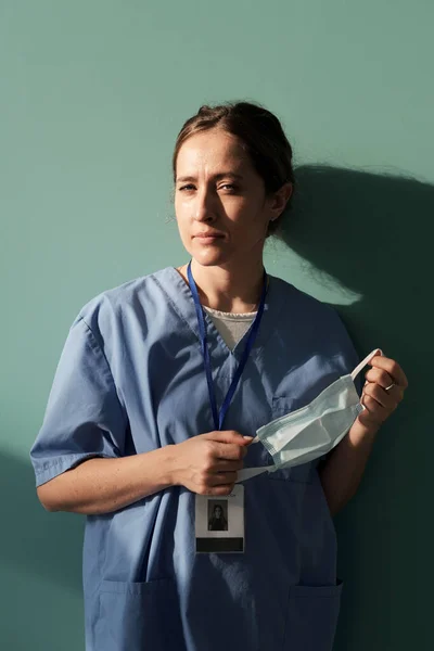 Young female assistant or physician in blue uniform holding protective mask in hands while standing by wall of hospital ward