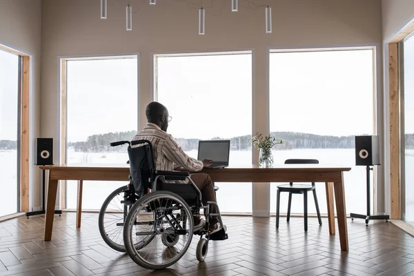 African American man with disability working in front of laptop while sitting by table in front of lapge window in living room