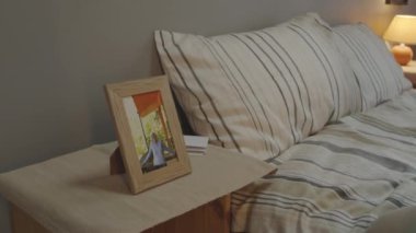 Depressed white-haired Caucasian man with mustache sitting on bed in his bedroom early in morning, looking at picture of wife in frame and thinking