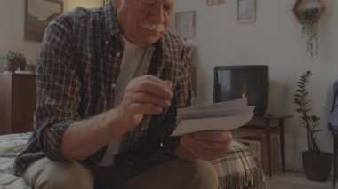 Waist-up of sad white-haired Caucasian man with mustache sitting in his bedroom early in morning, looking at old pictures and thinking