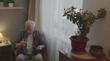 Medium of senior Caucasian man with mustache sitting in his room by window early in morning, taking pill, drinking from glass, then watering plant with same water