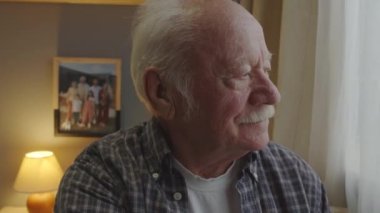 Shoulder-up of positive aged Caucasian man with mustache sitting in his room early in morning, looking out window and smiling