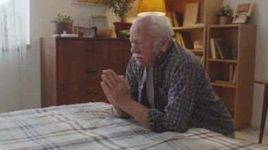 Medium of retired Caucasian man with white mustache praying in his room, kneeling by bed in evening