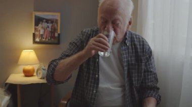 Waist-up of senior Caucasian man with mustache sitting in his room early in morning, taking pill, drinking water from glass, looking out window