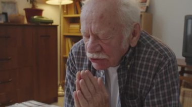 Medium close-up of retired Caucasian man with white mustache praying in his room, kneeling by bed in evening