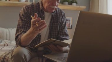 Tilting up of energetic senior Caucasian man sitting in his bedroom at daytime, video calling via portable computer, pensioner taking online consultation and writing in notebook