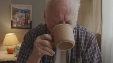 Chest-up of upset senior Caucasian man sitting indoors in morning, drinking coffee, looking out window