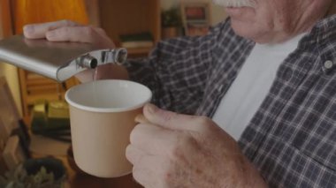 Close up of elderly Caucasian man with mustache pouring alcohol from flask into mug with coffee at home in morning, drinking