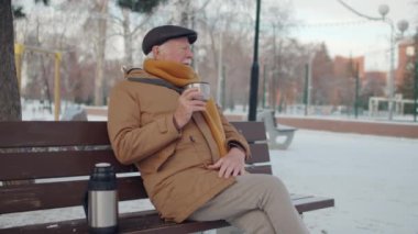 Medium long of senior Caucasian man wearing flat cap, jacket and scarf, sitting on bench in park in winter, drinking hot tea from vacuum flask