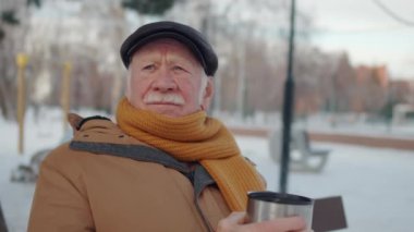 Chest up of senior Caucasian man wearing flat cap, jacket and scarf, sitting in park in winter, drinking hot tea