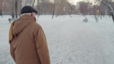 Steadicam rear of senior Caucasian man wearing flat cap, jacket and scarf, walking in park in winter