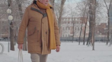 Tilting up of senior Caucasian man wearing flat cap, jacket and scarf, carrying net bag with fruit, walking in park in winter, reading text message on smartphone and smiling