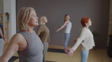 Side view of mature Caucasian people working out, stretching back and chest with female Biracial yoga instructor in class