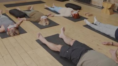 Tracking of aged women and men lying on backs on floor in modern yoga studio, meditating, young female fitness instructor walking around and talking