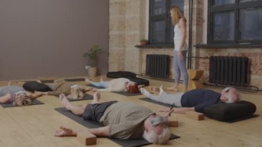 Long shot of senior women and men lying on backs on floor in modern yoga studio, meditating, young female fitness instructor walking and talking