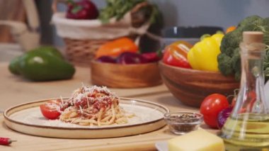 Close up tracking shot of plate with delicious spaghetti bolognese served with grated cheese on top and fresh tomato on side on kitchen table with various food ingredients