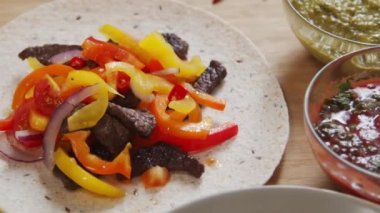 Close up tracking shot of flour tortilla with delicious beef and veggies filling on it and bowls with guacamole and salsa sauces for taco on kitchen table