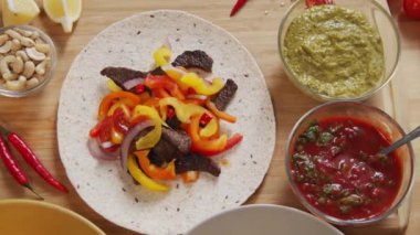 Top down shot of flour tortilla with beef and veggies on it, bowls with guacamole and salsa sauces and food ingredients for tacos on wooden table