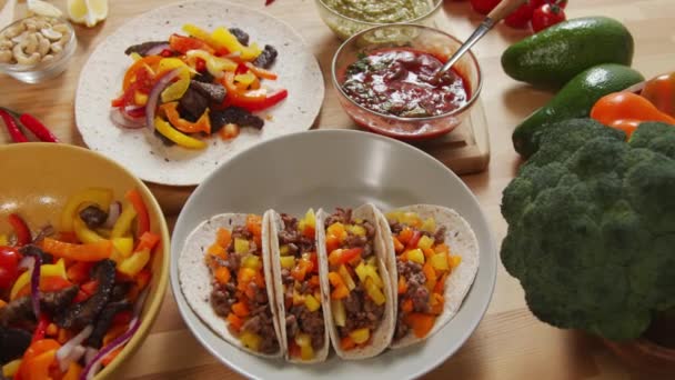 メキシコのタコスとプレートの高角度追跡ショット 木製のテーブルの上に調理された牛肉ストリップと野菜とボウル トルティーヤ ソースや食品成分 — ストック動画