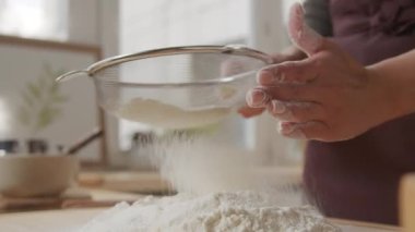 Close up view of hands of housewife sifting flour while cooking food in kitchen at home