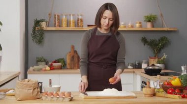 Woman in apron cracking egg with knife and adding it to flour while preparing dough at kitchen table