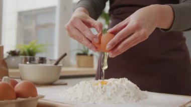 Cropped shot of unrecognizable woman in apron adding egg to flour pile while preparing dough at kitchen table