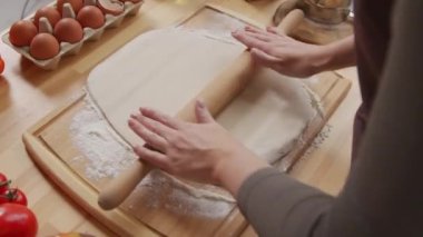 Over the shoulder shot of woman rolling pasta dough on wooden board at kitchen table