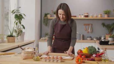 Caucasian woman in apron standing by kitchen table and rolling dough while cooking at home