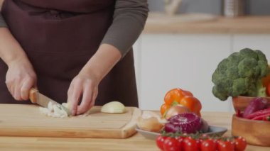 Close up tracking shot of unrecognizable woman in apron chopping onion with knife on wooden cutting board at kitchen table