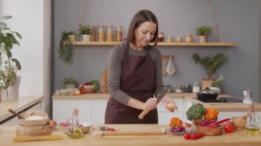 Medium shot of woman in apron peeling onion with knife while cooking dinner in kitchen at home