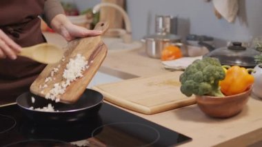 Cropped shot of woman in apron putting chopped onion on hot pan and frying it while cooking in kitchen at home