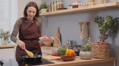 Housewife in apron putting ground meat from bowl in hot frying pan while cooking food in kitchen at home