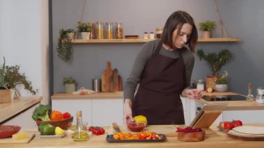 Woman in apron preparing food ingredients and watching video recipe on digital tablet while cooking dinner at home