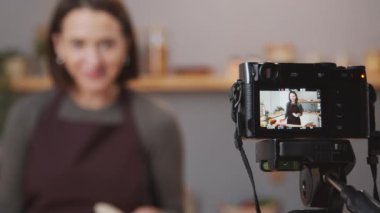 Selective focus shot of professional camera on tripod filming female food blogger telling dinner recipe in kitchen
