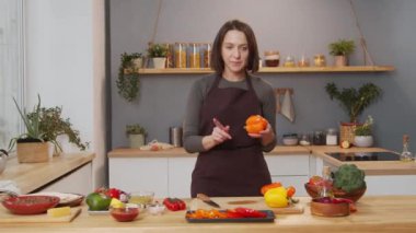 Female food blogger in apron standing in kitchen, showing ingredients and telling recipe on camera