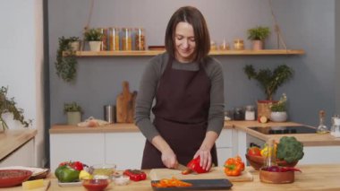 Woman in apron cutting fresh bell pepper while cooking dinner in kitchen