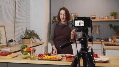 Woman in apron showing food ingredients and telling about them while filming culinary vlog with professional camera in kitchen