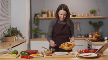 Caucasian woman in apron putting meat and veggies filling on tortilla while preparing tacos in kitchen at home