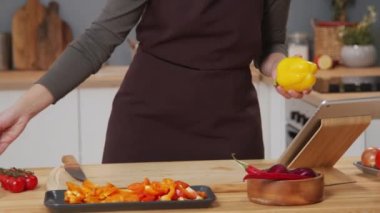 Cropped shot of woman searching food recipe on the Internet on digital tablet, then taking ingredients and cutting bell pepper while cooking meal at kitchen table