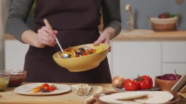 Cropped tracking shot of woman putting beef strips with pepper slices on flour tortilla while making mexican tacos at kitchen table