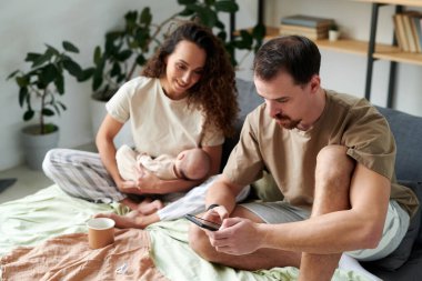 Serious young man texting in smartphone while sitting on double bed next to his wife with baby son looking at screen of gadget