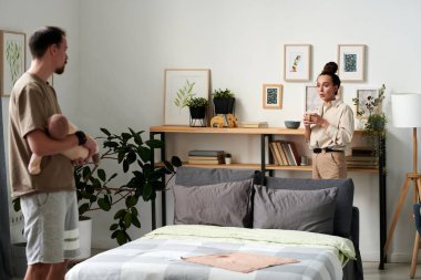 Young woman with cup of coffee standing by doubel bed in front of her husband holding sleeping baby son on hands