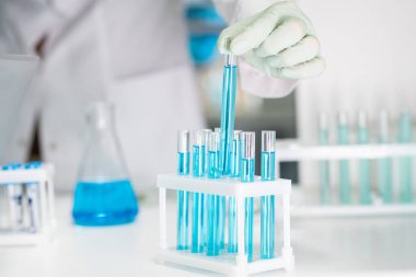 Gloved hand of female chemist or biotechnologist taking one of many flasks containing blue liquid chemical substance in laboratory