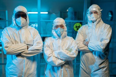 Group of contemporary interracial researchers in protective workwear crossing arms by chest while standing against laboratory