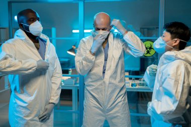 Three interracial scientists putting on protective coveralls and respirators while going to work with biohazard substances in laboratory