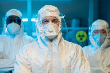 Mature male leader of research group in white protective coveralls, facial screen and respirator standing against his two colleagues